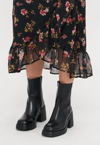 Sorte lær ankler støvler med en kraftig høy hæl, med glidelås på siden og en jevn tekstur. Kjolen har et blomsterdesign med volanger i kanten.