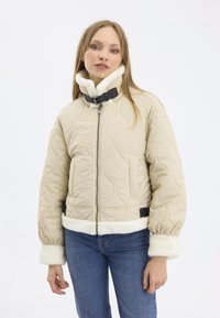 Veste matelassée beige avec un col haut, ornée d'une garniture en fausse fourrure blanche et de accents en boucle noire. Comprend des poches latérales et des poignets élastiques.