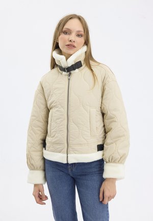 Beige quiltet jakke med høy krage som har hvit kunstpels og svarte spenne detaljer. Har sidelommer og elastiske mansjetter.