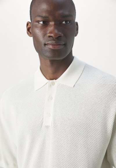 Polo shirt en mesh blanc avec un col et un rabat à trois boutons. Il présente un tissu texturé et un design amincissant.