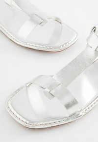 Silverfärgade metallicsandaler med fyrkantig tådesign. Har detaljer med korsband, slät lädertextur och kontrasterande vit söm.