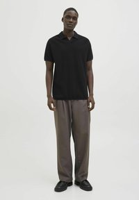 Polo shirt nero a maniche corte con una consistenza morbida, abbinato a pantaloni grigi larghi e scarpe nere, in piedi davanti a uno sfondo neutro.