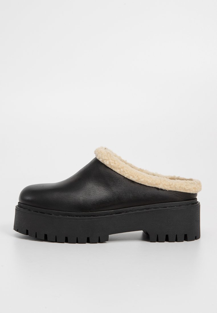 Sabots en cuir noir avec un col en shearling beige, semelle épaisse en caoutchouc noir avec motif, bout arrondi et design facile à enfiler.