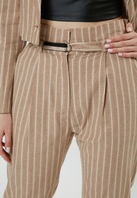 Pantalones a rayas marrón claro con una textura sutil, que incluyen un cinturón con un anillo metálico brillante y un ligero detalle de pliegue en la cintura.