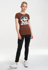 Brown fitted t-shirt with short sleeves featuring a graphic design of a superhero and text "Girls Will Save the World" in white.