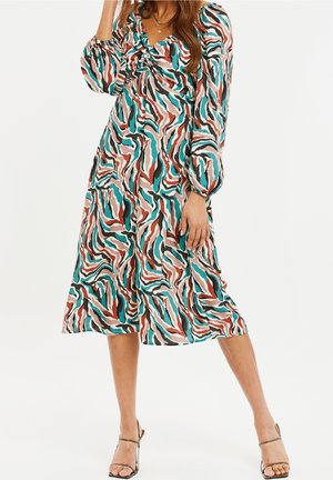 Femme portant une robe à manches longues, longueur genou, avec des motifs abstraits ondulés en bleu sarcelle, rouille, noir et blanc, associée à des sandales à talons hauts à brides.