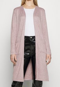 Lång rosa cardigan av mjukt material, med en avslappnad passform, öppen front och två framfickor. Kombinerad med blanka svarta byxor.