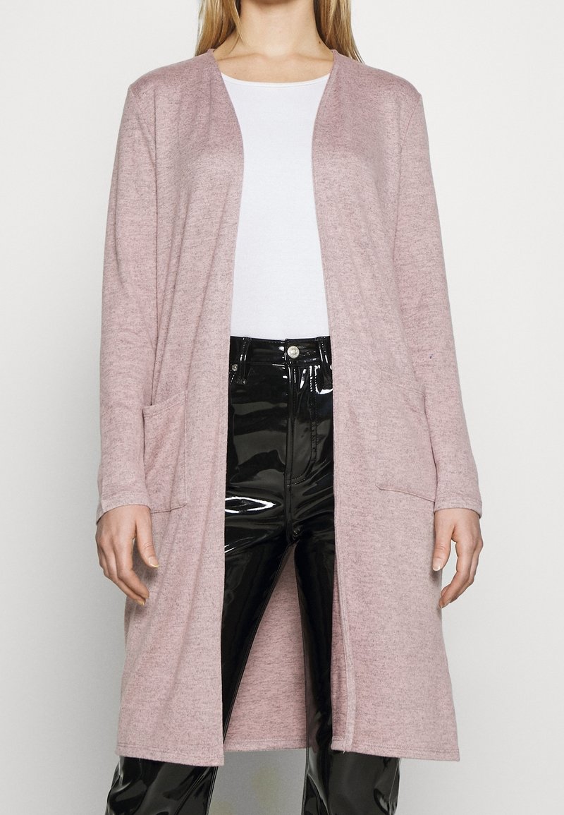 Lång rosa cardigan av mjukt material, med en avslappnad passform, öppen front och två framfickor. Kombinerad med blanka svarta byxor.