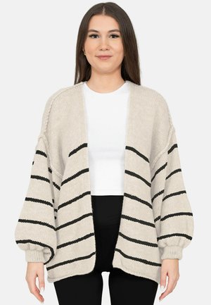 Jeune femme portant un cardigan beige en maille avec des rayures noires horizontales, sur un haut blanc et un pantalon noir, debout devant un fond uni.