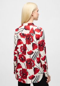 Langarmblouse met bloemenpatroon in rood en zwart op een crèmekleurige achtergrond. Heeft een lichte glans, een kraag en een rechte zoom.