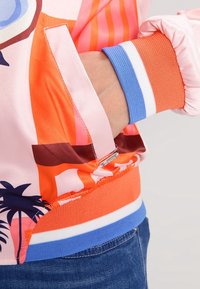 Giacca bomber colorata con una finitura lucida simile alla satin. Presenta un motivo astratto di palme, polsini a costine in contrasto e una tasca con zip.