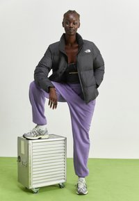Giacca a piumino nera con collo alto, abbinata a pantaloni sportivi viola. Il modello sta in piedi su una valigia argentata a coste, indossando sneakers grigie.