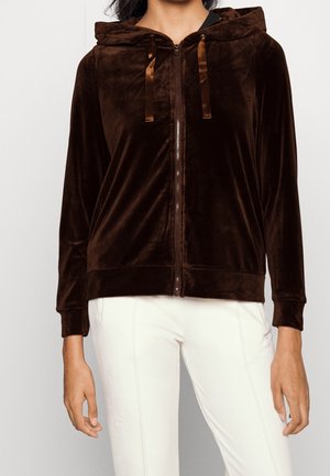 Kobieta ubrana w ciemnobrązową welurową bluzę z kapturem zapinaną na zamek z satynowymi troczkami oraz białe dopasowane spodnie, pokazana od ramion do bioder.