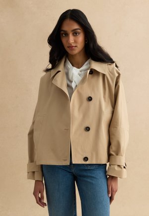 Vrouw met lang donker haar, gekleed in een beige jasje met knopen over een wit blouse en een blauwe spijkerbroek, staand tegen een effen beige achtergrond.