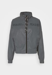 Grijze cropped jas van lichtgewicht materiaal met een hoge kraag, voorzien van het opgelegde "KARL KANI"-logo en elastische boorden bij de mouwen en zoom. Ritssluiting aan de voorkant.