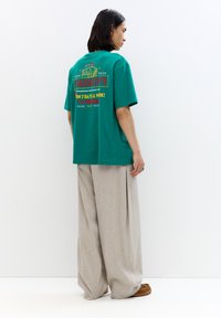 Tricou din bumbac verde, cu un text colorat și un design grafic pe spate, asortat cu pantaloni largi bej și încălțăminte maro.