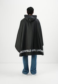 Persoon in een zwart, met een capuchon, regenponcho met de tekst "KARL LAGERFELD" nabij de onderkant, blauwe spijkerbroek en witte sneakers, met de rug naar de kijker gekeerd.