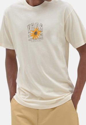 Beżowy T-shirt Vans z żółtą grafiką kwiatu, napis „Vans 1966 Off The Wall Original”, noszony z jasnobrązowymi spodniami i rękami w kieszeniach.