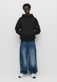 Black hoodie with drawstrings, oversized fit. Paired with loose-fitting denim jeans, faded blue with a distressed look, and white sneakers.