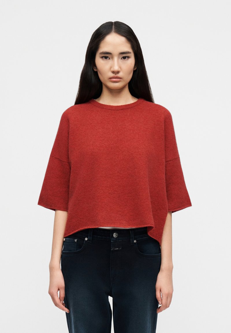 Κόκκινο, υπερμεγέθες πλεκτό πουλόβερ με κοντά μανίκια και στρογγυλή λαιμόκοψη, σε συνδυασμό με σκουρόχρωμο τζιν παντελόνι. Απαλή υφή, casual σχεδιασμός.