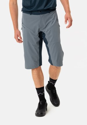 Shorts gris à plusieurs panneaux avec des accents bleu foncé, comportant des poches zippées sur les côtés et un design de taille confortable. Portés avec des chaussures et des chaussettes noires.