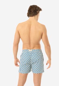 Mann, der mit dem Rücken zur Kamera steht, trägt blau-weiße, geometrisch gemusterte Badeshorts vor einem schlichten weißen Hintergrund.