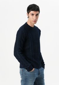 Navyblauwe gebreide trui met een gestructureerd kabelpatroon, ronde halslijn en geribde manchetten. Gemaakt met lichtblauwe jeans.
