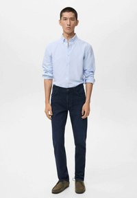 Un hombre está de pie delante, vistiendo una camisa de botones azul claro, jeans azul oscuro y zapatos casuales marrones, contra un fondo blanco liso.
