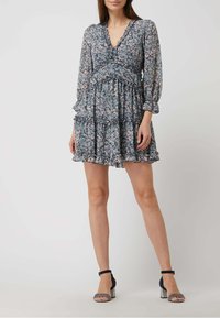 Femme portant une robe florale courte à manches longues avec des volants et des sandales à talons hauts à bride de cheville sur un fond uni clair.