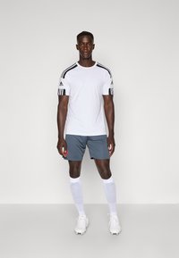 Hvid sports t-shirt med sorte striber på skuldrene, parret med grå shorts, hvide knæstrømper og hvide fodboldstøvler. Minimalistisk design.