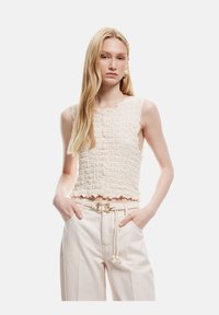 Top sin mangas de color crema, texturizado, con un dobladillo festoneado, combinado con pantalones beige claros que tienen un cinturón y borlas decorativas.