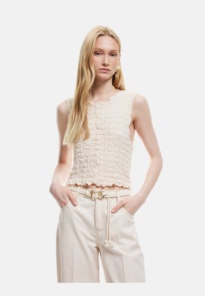 Top senza maniche color crema, texture, con orlo scallop, abbinato a pantaloni beige chiaro con cintura e frange decorative.