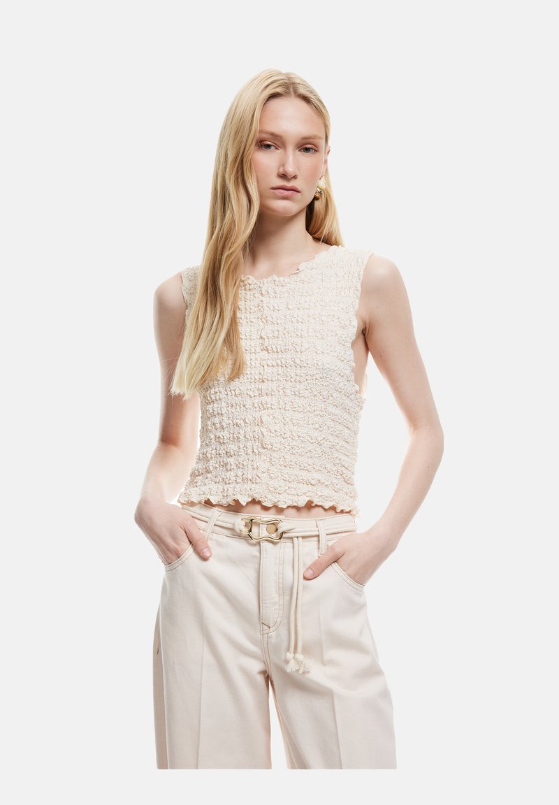 Top sin mangas de color crema, texturizado, con un dobladillo festoneado, combinado con pantalones beige claros que tienen un cinturón y borlas decorativas.