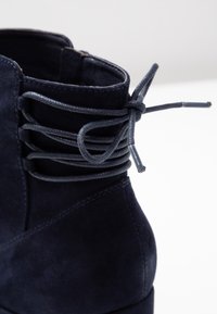 Granatowe zamszowe botki sięgające kostki z zaokrąglonym noskiem i krzyżowym sznurowaniem w okolicy kołnierza. Gładka tekstura z subtelnym połyskiem.
