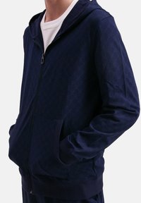 Uomo che indossa una felpa blu navy con zip, caratterizzata da una lieve trama decorativa, con le mani infilate nelle tasche anteriori, sopra una maglietta bianca a girocollo.