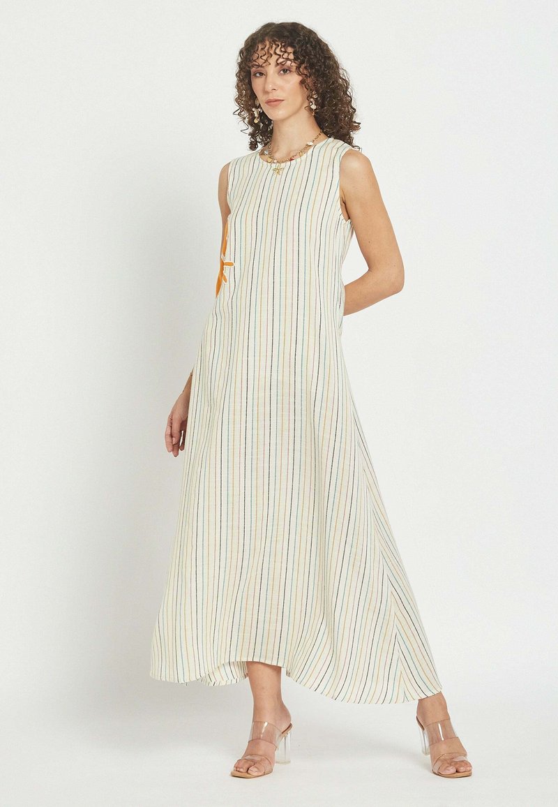 Mujer con cabello rizado que lleva un vestido blanco sin mangas hasta los tobillos con finas rayas verticales y sandalias con tacón transparente.