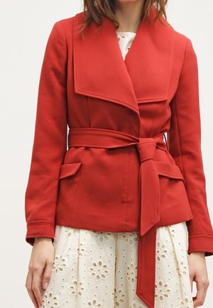 Femme portant une veste rouge ceinturée sur une robe blanche à œillets, debout devant un fond clair uni.