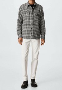 Homme portant une chemise à carreaux pied-de-poule noire et blanche, un pantalon blanc et des bottes en cuir noires, debout devant un fond uni.