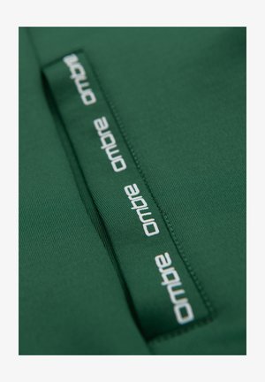 Tissu vert avec une couture cousue et une bande de tissu présentant un texte blanc « ombre » répété dans une police moderne.