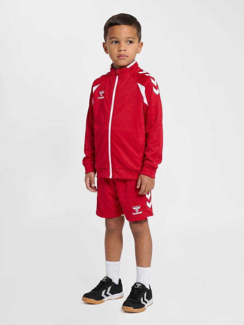 Garçon debout portant une veste de sport rouge et un short avec des accents blancs, des chaussettes blanches et des chaussures de sport noires sur un fond blanc.