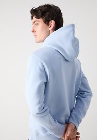 Light blue hoodie made of soft fabric, featuring a large adjustable hood, ribbed cuffs, and a relaxed fit. Back view shows stitching details.
