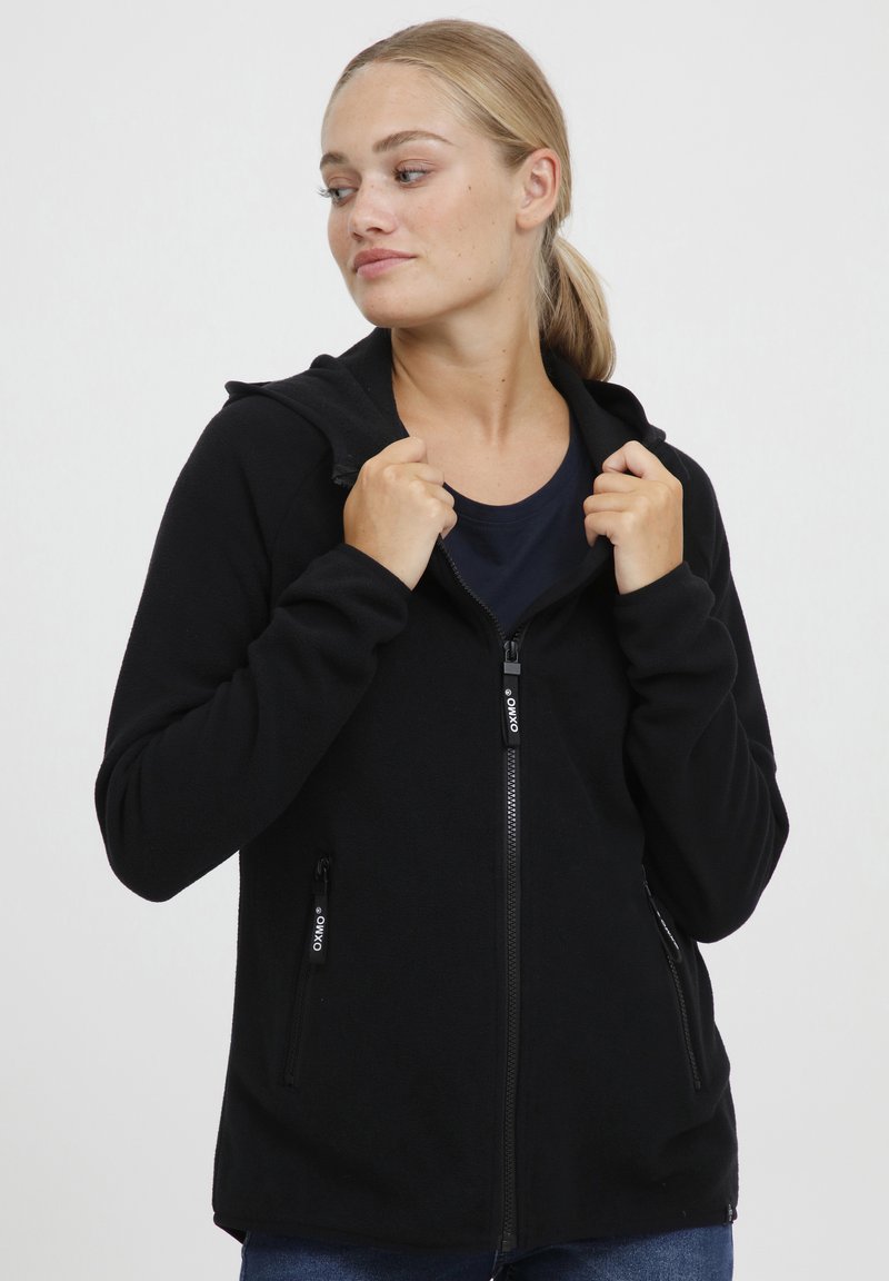 Svart fleece hettegenser med glidelås foran og to sidelommer, med hette og glatt tekstur. Minimal merkevare på glidelåsen.
