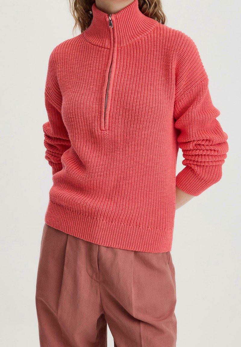 Korallinvärinen neulepaita, jossa korkea vetoketjullinen kaulus, resorikuvio ja pitkät hihat. Yhdistetty väljiin vaaleanpunaisiin housuihin.