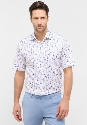 Man draagt een wit overhemd met korte mouwen en een patroon van blauwe zeilboten, gecombineerd met lichtblauwe broek, tegen een effen achtergrond.