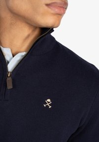 Sudadera azul marino con cremallera hecha de tejido de punto, con un cuello azul claro y un pequeño emblema dorado en el pecho. La cremallera tiene un tirador dorado.