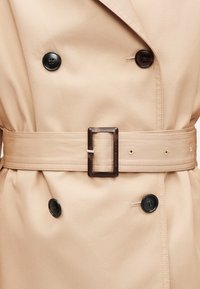 Μπεζ παλτό τύπου τραντσκότ με διπλό πέτο, μαύρα κουμπιά και υφασμάτινη ζώνη με ορθογώνια καφέ αγκράφα.