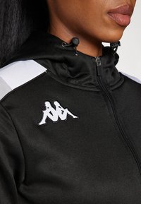 Svart zip-jacka med hög krage, vita axelaccenter och en broderad vit logotyp på bröstet. Slät tygstruktur.