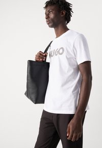 Svart lær tote bag med en strukturert form og en enkel stropp, kombinert med en hvit bomulls-t-skjorte med metallisk logo-trykk.