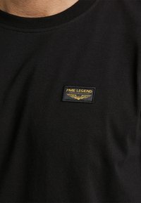 Schwarzes Shirt mit einem kleinen rechteckigen PME Legend American Classic Logo-Patch, das einen gelben Adler auf der Brust zeigt.