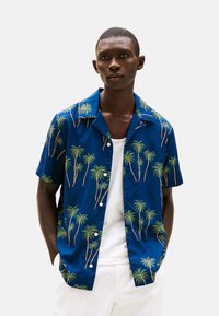 Camisa de manga corta, con botones, de tela azul con un estampado de palmeras en verde y marrón. Textura suave con diseño de cuello.